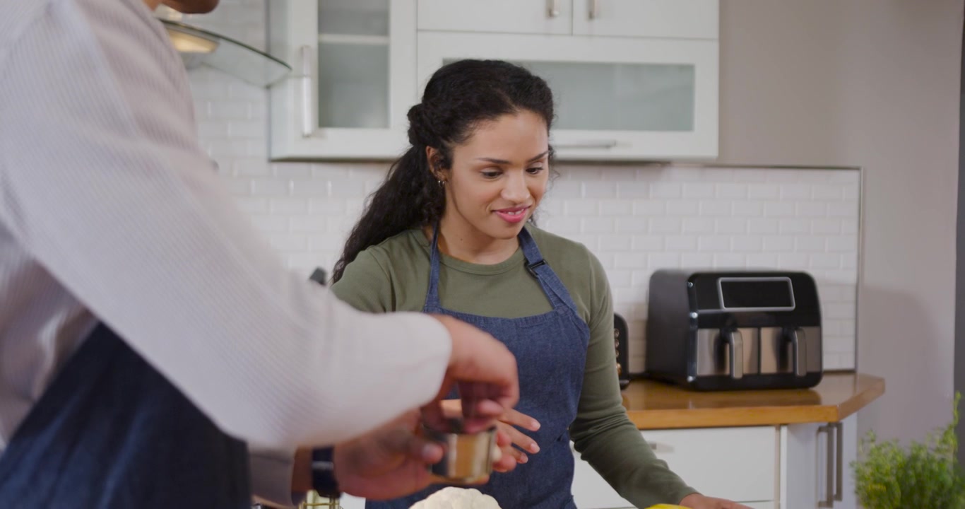 Casal afro-americano usando aventais temperando uma bandeja na bancada da cozinha enquanto o homem oferece xícaras para assar.