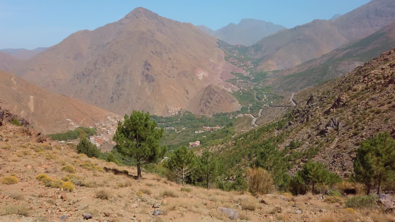 Vista sulla Valle di Imlil nelle Alte montagne dell'Atlante in Marocco, che mostra pendii scoscesi, pini sparsi e piccoli villaggi berberi lungo una strada di montagna tortuosa nella pittoresca regione del Monte Toubkal.