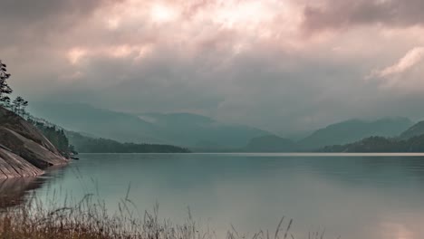 Durchdringende-Sonnenstrahlen-Scheinen-Durch-Die-Stürmischen-Wolken,-Die-über-Dem-See-Mit-Felsigen-Ufern-Und-Bergen-In-Einem-Zeitraffervideo-Wirbeln