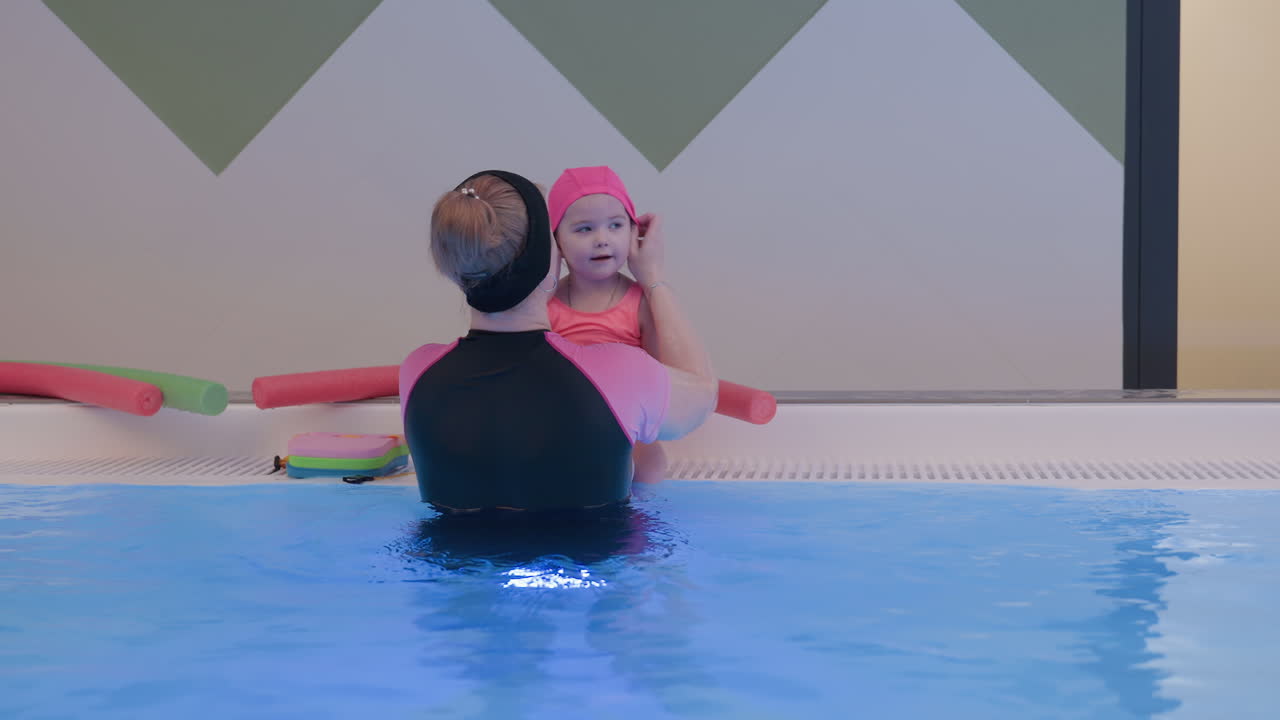 Lady inside pool adjusting toddler head cap, gentle hands fixing fit, child smiling, blue water shimmer, safe supervised moment, warm daylight, family swim practice, soft blur, calm playful mood