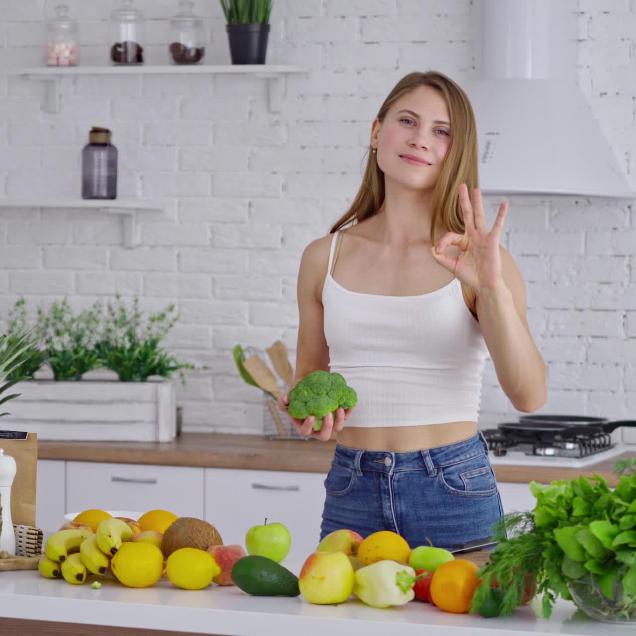 Cheerful female with broccoli at home. Sportive young woman choosing healthy diet full of minerals and vitamins in the kitchen. Girl standing near fresh fruit and vegetables.