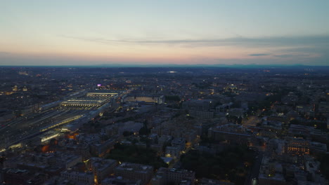 Vista-Panorámica-Aérea-De-La-Ciudad-Nocturna-Y-El-Cielo-Crepuscular-De-Color-En-Segundo-Plano.-Estación-Central-De-Tren-Ricamente-Iluminada.-Roma,-Italia