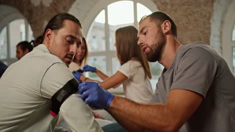 Close-up-shooting:-A-bearded-brunette-man-in-a-gray-T-shirt-puts-a-tourniquet-on-the-hand-of-a-brunette-man-with-long-hair-during-a-practical-medical-course