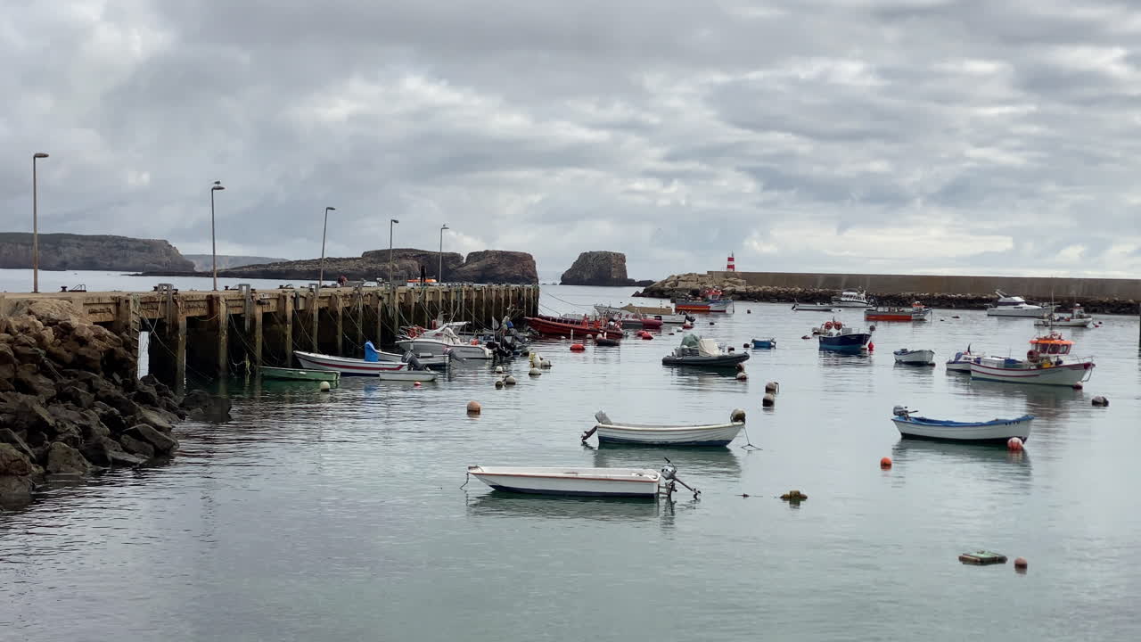 Fishing boats float calmly in Sagres harbor beneath golden cliffs and cloudy Atlantic skies