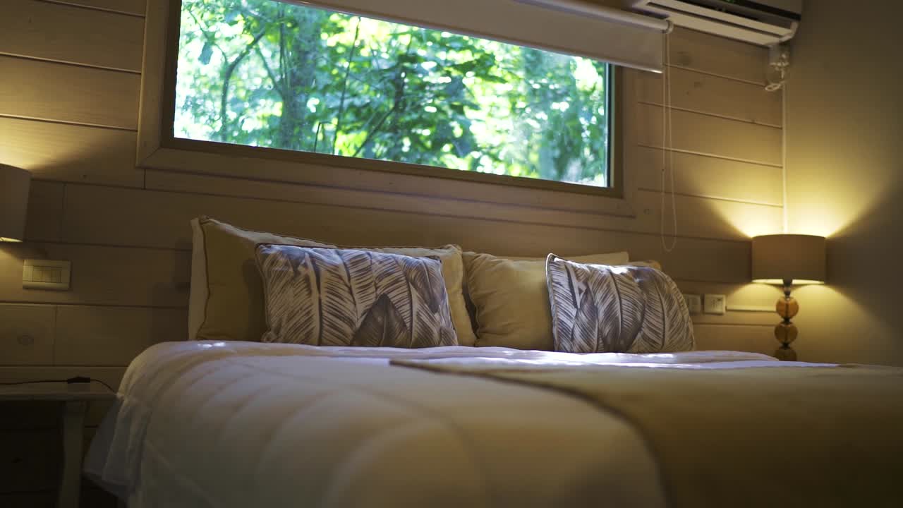 A slow-moving shot reveals a luxurious hotel room nestled in the jungle, featuring a large kingsize bed elegantly made up in a serene, tropical setting, blending nature and comfort.