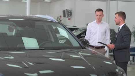 young guy standing near nice black sports car talking to dealer side view car business car sales technology and people concept happy man and car dealer with tablet pc in car showroom or showroom