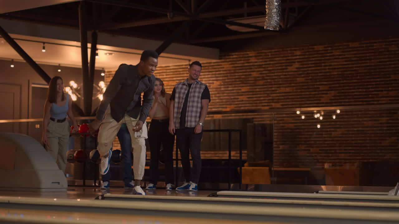 Un uomo afroamericano lancia una palla da bowling e fa strike con un solo lancio, abbraccia e festeggia con gli amici. Gruppo multietnico di amici che giocano a bowling