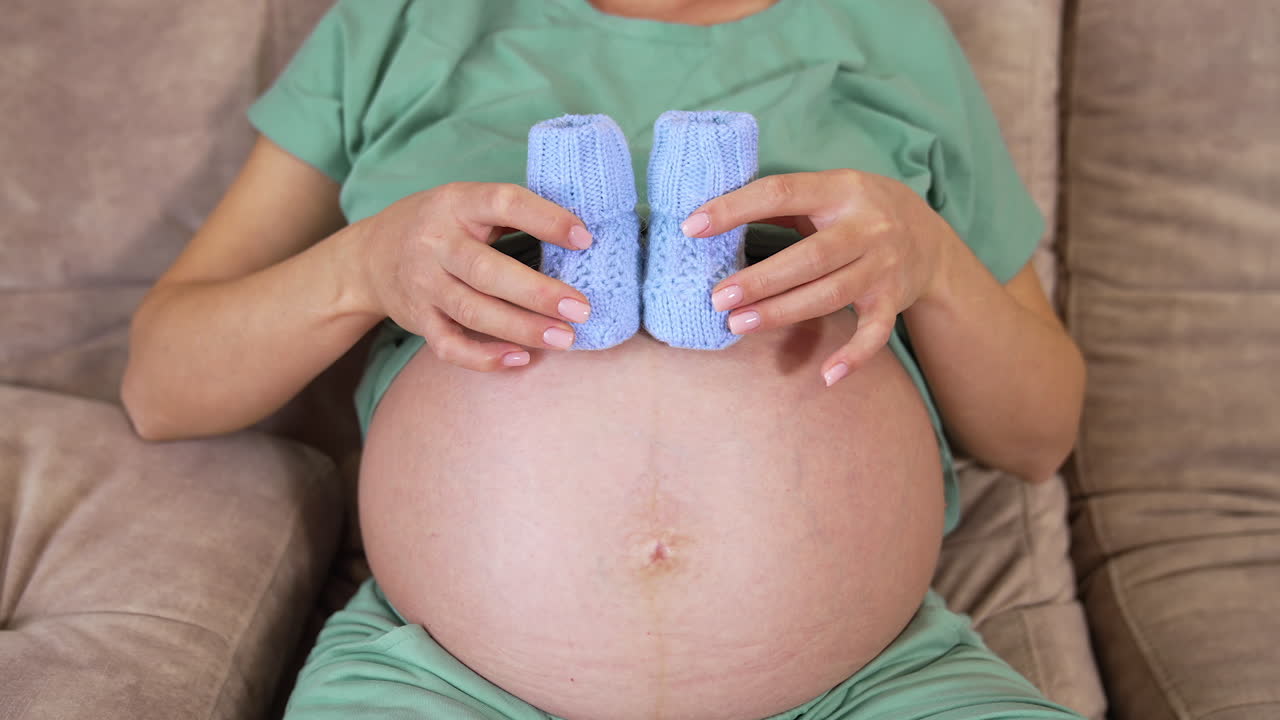 Knitted socks on a bare belly of a future mother. Pregnant woman laying on a sofa and shows little blue socks on a belly. Front view.