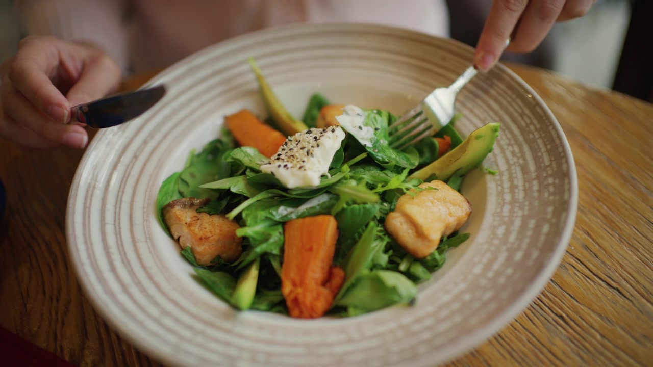 Primer plano de una mujer comiendo una ensalada colorida con hojas verdes, rodajas de aguacate, trozos de salmón, calabaza naranja asada en un tazón ancho a rayas