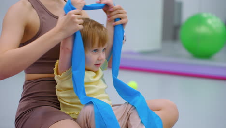 close-up of a little girl seated with her mother, both playfully holding and moving a ribbon back and forth during a workout session in a gym