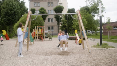 Front-view-of-little-girl-with-down-syndrome-wearing-hoodie-swinging-on-a-swing-in-the-park-on-a-windy-day.-Her-male-friend-pushing-her-while-her-female-friend-take-a-photo