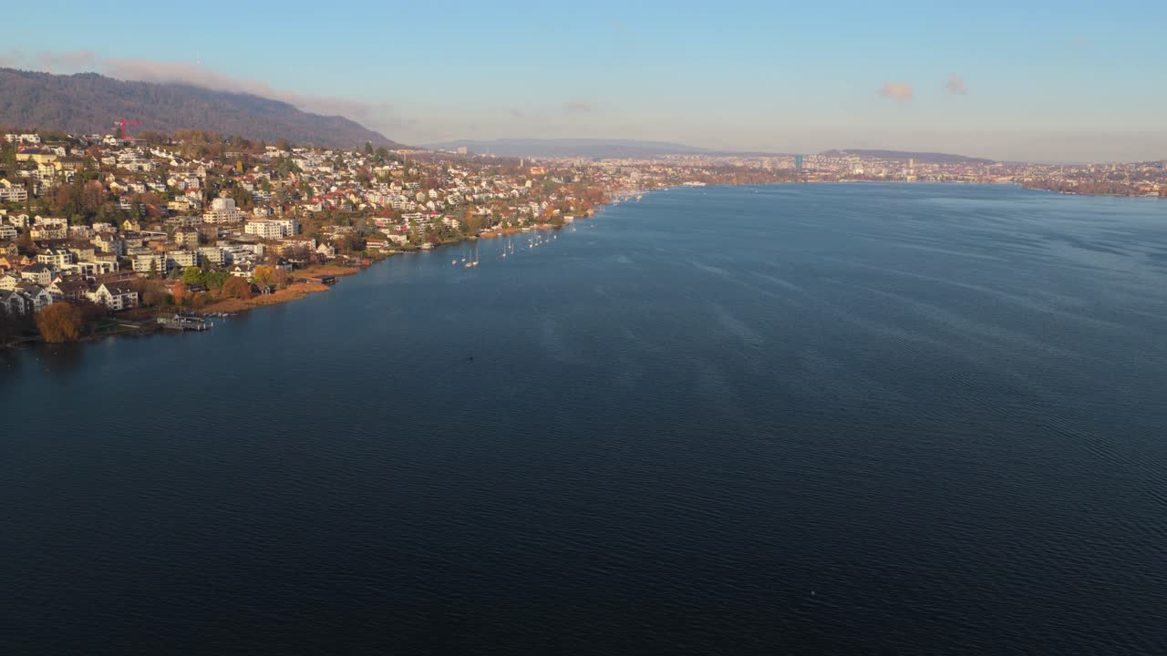 Drone riprende un'ampia vista aerea dal centro del lago di Zurigo. La chiara luce del giorno rivela la costa di Kilchberg e la città di Zurigo in lontananza, al di là delle calme acque blu