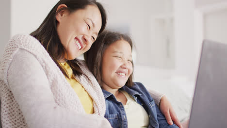 Video-of-happy-asian-mother-and-daughter-sitting-on-sofa-with-laptop