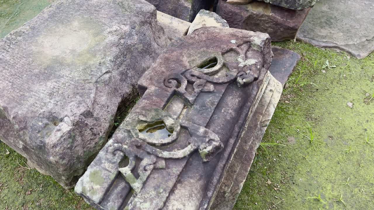 A static shot of a weathered stone block featuring faint ornamental carvings. Its moss-stained surface and worn edges speak of centuries of history, artistry, and natural erosion