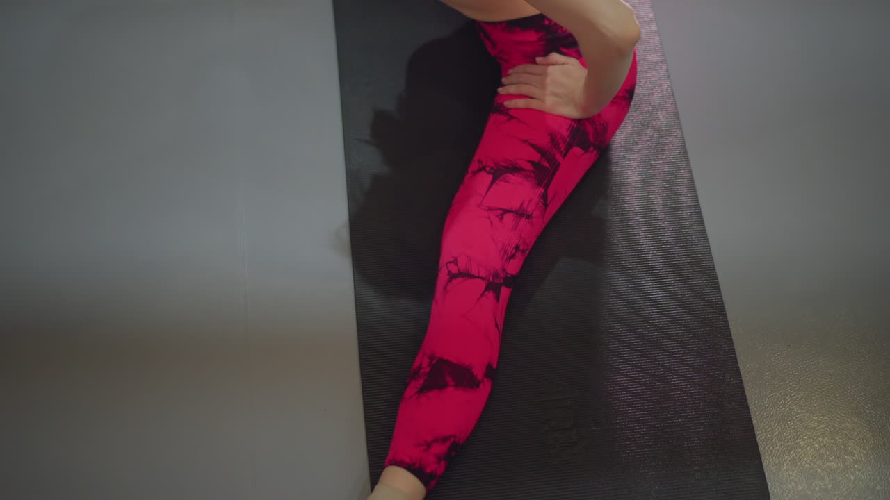 Donna asiatica che esegue stretching da seduta su tappetino, vista ravvicinata dall'alto che mostra i leggings rosa, respirazione concentrata e mobilità lenta per il recupero dei muscoli posteriori della coscia, con un'atmosfera da studentessa di yoga e cliente in riabilitazione, guida delicata dell'istruttore, calma.