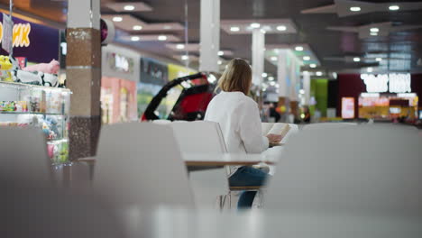 young woman reading a book in a mall, seated at a table with a soft focus on her surroundings, the background features vibrant lights, shops, and people moving