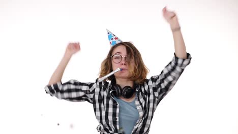 Cheerful,-excited-young-woman-in-plaid-shirt-and-headphones-on-neck-in-birthday-colorful-hat-blow-in-pipe-and-dancing,-falling-confetti-isolated-over-white-background-in-studio.-People-sincere-emotions,-holiday-lifestyle-concept.-Celebrating-party-day