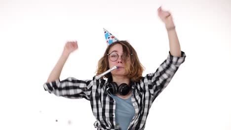 cheerful, excited young woman in plaid shirt and headphones on neck in birthday colorful hat blow in pipe and dancing, falling confetti isolated over white background in studio. people sincere emotions, holiday lifestyle concept. celebrating party day