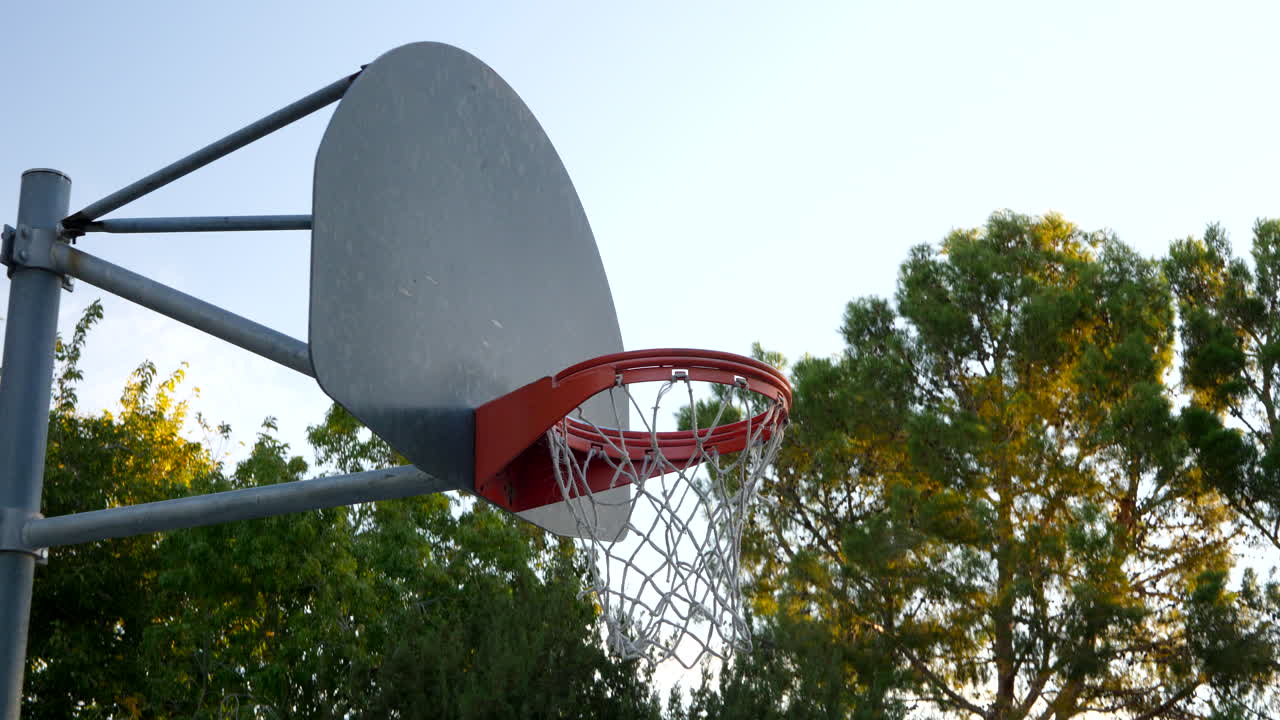 caminando hacia un aro de baloncesto con un tablero de metal y un borde naranja en una cancha de parque vacía al amanecer