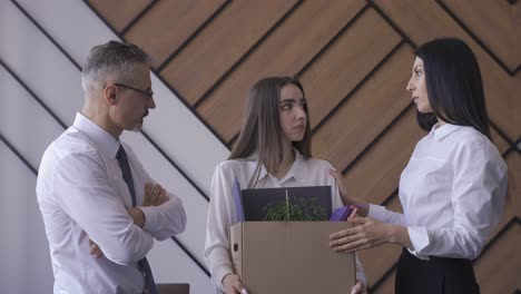 dos compañeros de trabajo consolando a otra empleada por su renuncia. la empleada sale de la oficina sosteniendo una caja con sus objetos personales