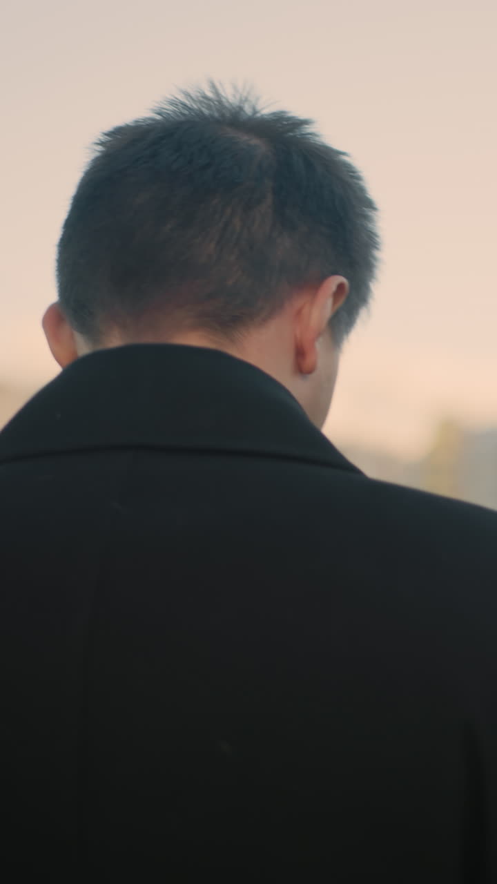 검은 코트를 입은 개인이 차를 마시며 도시 환경에서 사려 깊게 앞을 바라보고있는 뒷면, 부드러운 빛 아래 자동차와 높은 건물로 흐릿한 배경을 특징으로합니다.