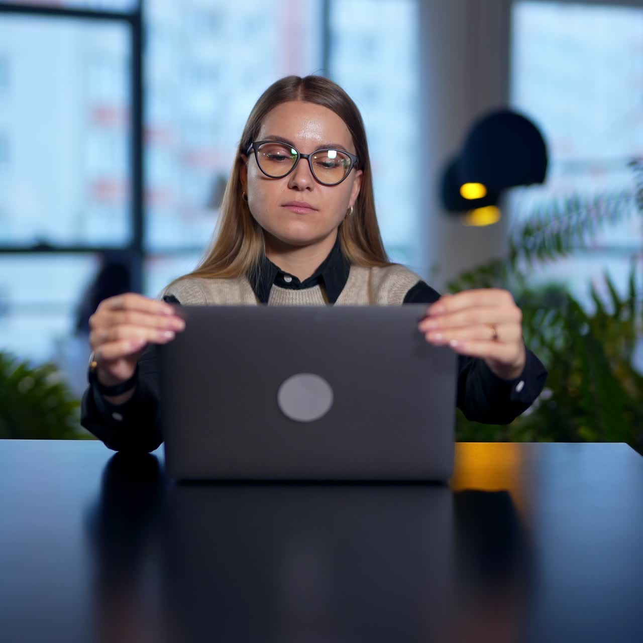 Lady in glasses opens laptop, prints something and closes it. Long-haired woman puts off glasses and lays her head on hands. Blurred backdrop