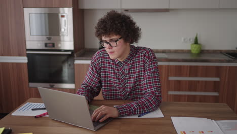 a curly man with a serious look works at a laptop sitting in a modern kitchen. young man freelancer student using laptop studying online working from home in internet