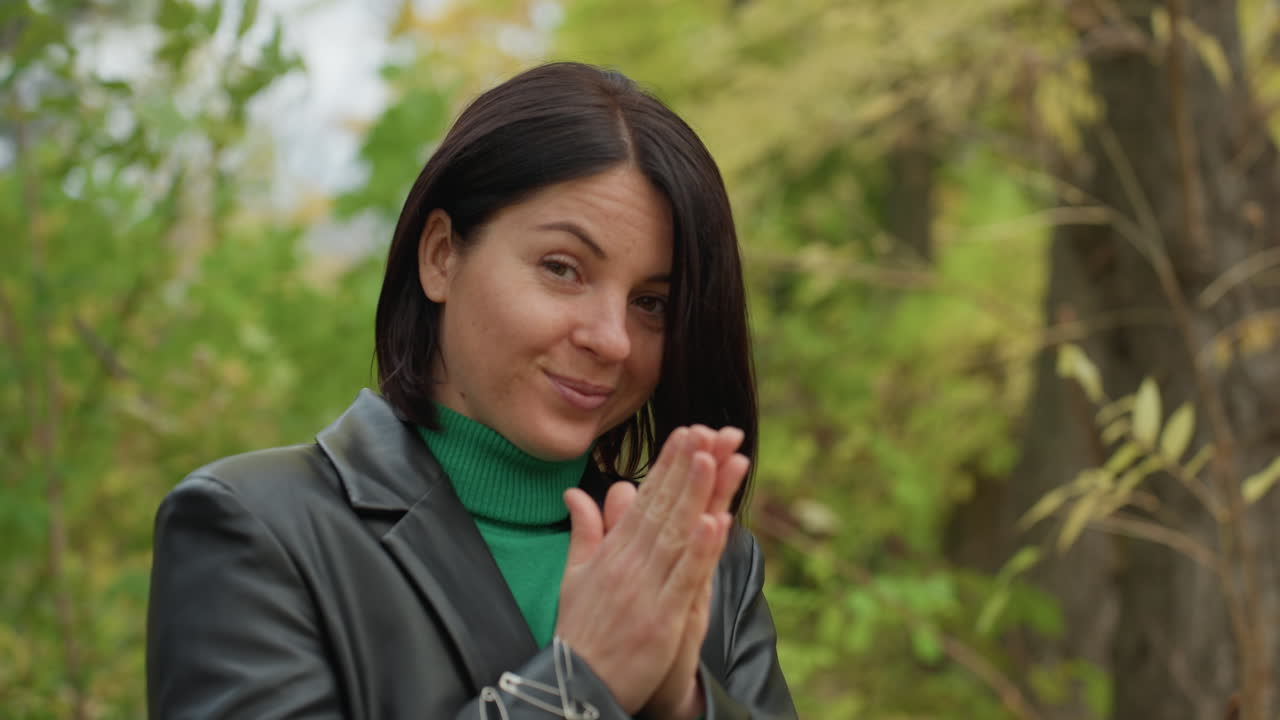 woman leather jacket meditating in forest, clasping hands in prayerlike clap, soft smile, green turtleneck visible, autumn leaves and tree trunks in background, leather studs and bracelet detail