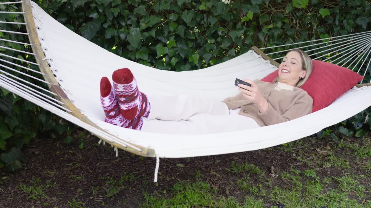 Donna che si sistema sull'amaca in giardino e avvicina la carta allo smartphone per pagare, festeggiando e rilassandosi