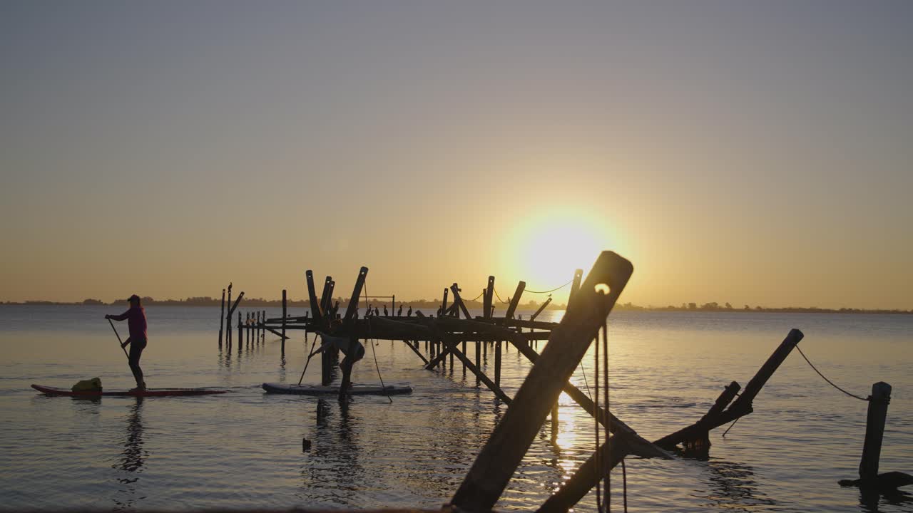 People Paddleboarding on a Calm Lake at Sunset with a Broken Pier