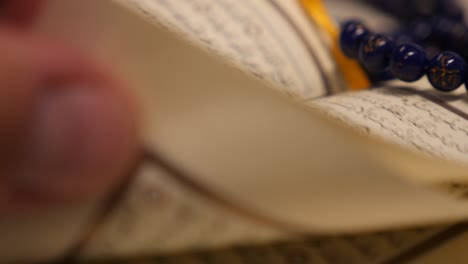 footage of a quran and a prayers bead near it, the shot is moving in a half circle and a person leafs through the quran