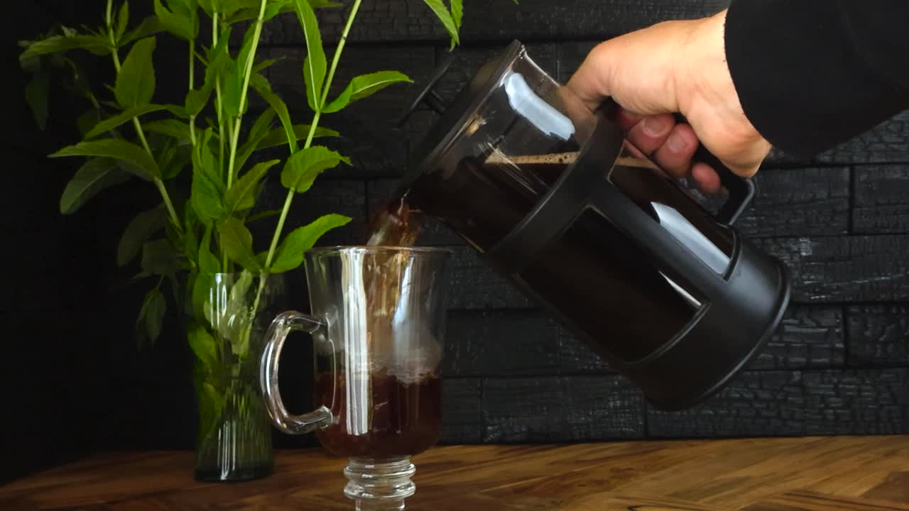 Slow motion footage of a caucasian man pouring coffee from a french press coffee can into a mug or a glass jar that is on an olive wood kitchen table and Yakisugi burnt wood panels at back. Peppermint