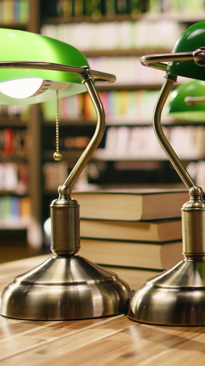 Vertical Video Stack of books placed on wooden library desk beneath green lamps