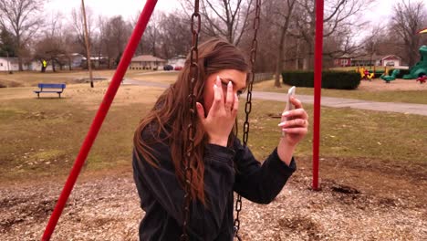 4k footage of a gorgeous college teenager sitting on a swing set using her cell phone as a mirror to check her makeup and hair