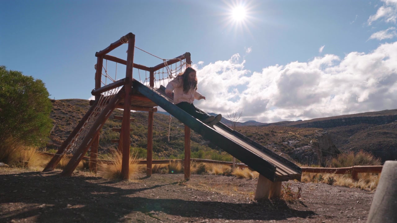 Scivolo da parco giochi rustico in un ambiente montano arido con luce solare intensa e nuvole drammatiche.