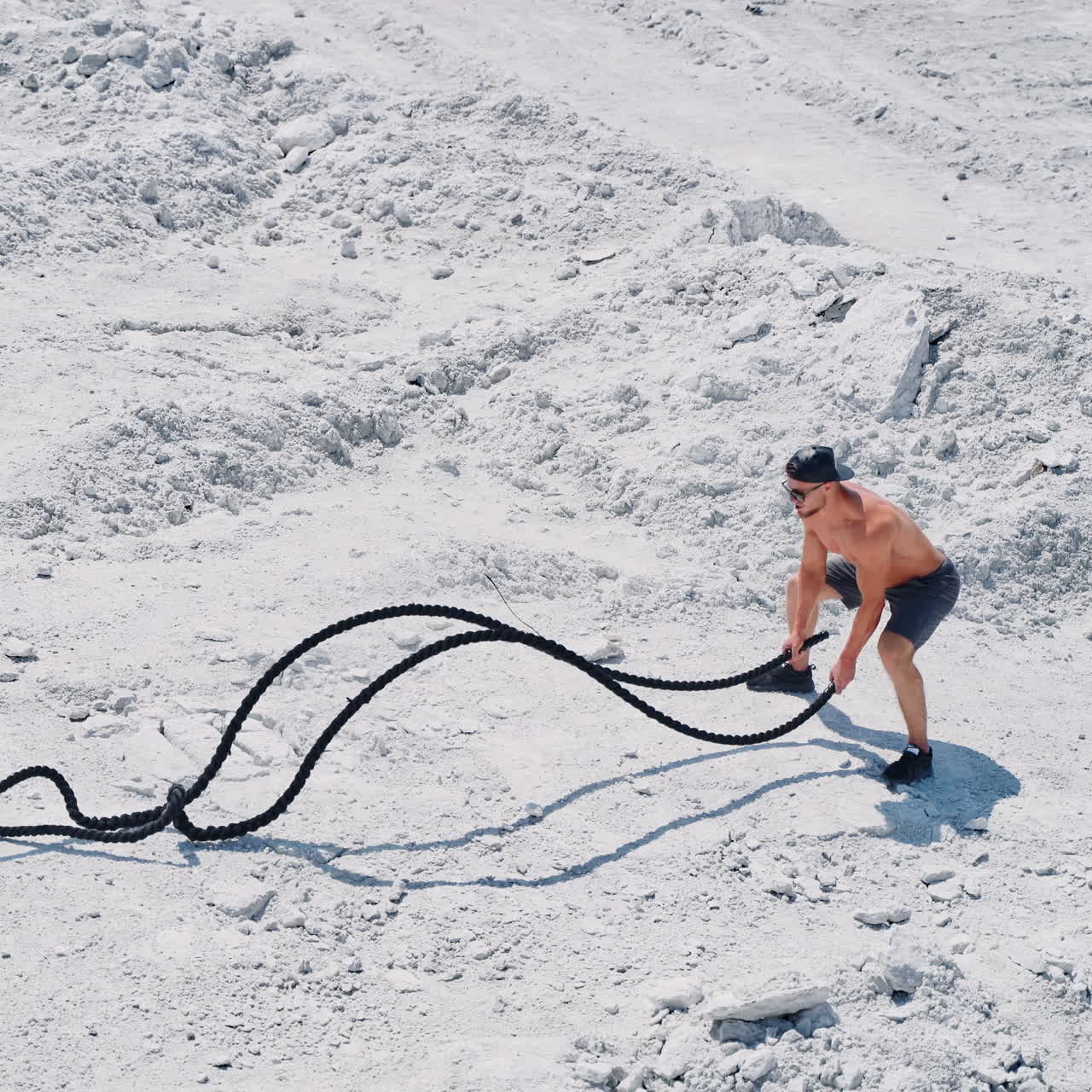 Strong young man doing exercises with a battle rope in sunny day. View from above on a muscular man training with cable on white ground outside. Concept of professional training.