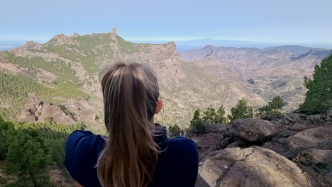 unrecognizable blonde girl observes the view and the roque nublo from the top of a mountain with her back to the viewer