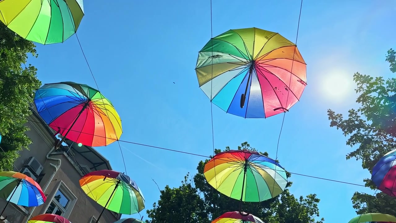 Farverige paraplyer hænger over en grøn park på en solrig dag under en lokal fest med lyse flag, der blafrer.