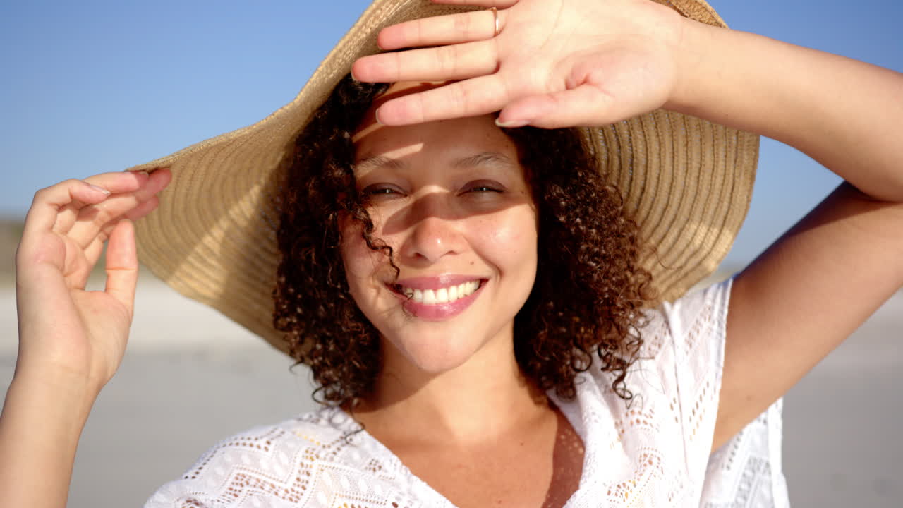 Una giovane donna birazziale con i capelli ricci sorride su una spiaggia assolata, proteggendosi gli occhi con la mano