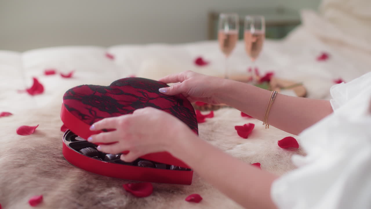 Woman unboxing Valentine's Day chocolates with rose wine, bright bedroom setting