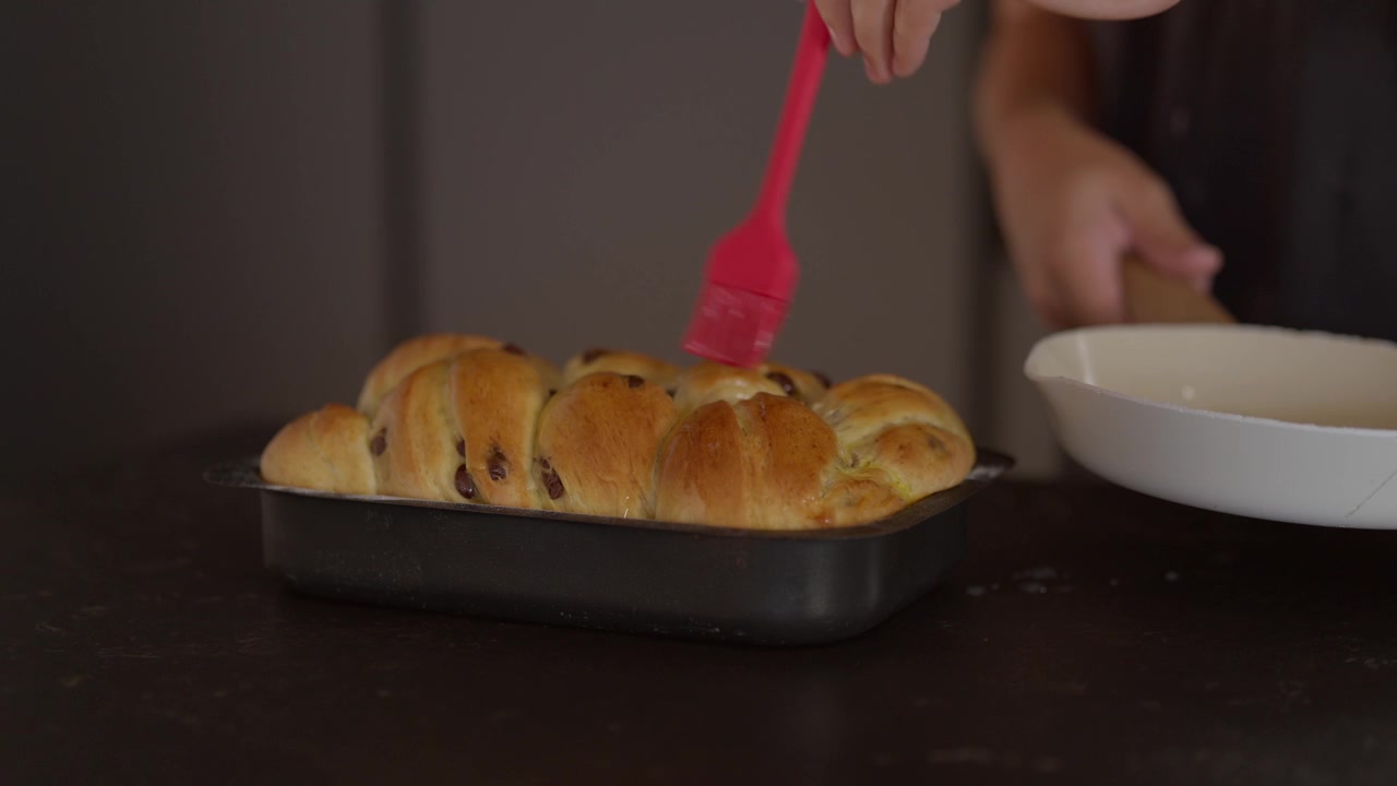 Hand brushing egg wash onto a freshly braided raisin bread loaf in a baking pan, preparing for baking. Homemade sweet bread