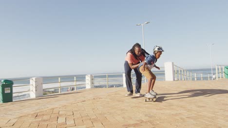 Video-Eines-Glücklichen-Afroamerikanischen-Vaters,-Der-Seinem-Sohn-Das-Skateboarden-Auf-Der-Promenade-Beibringt