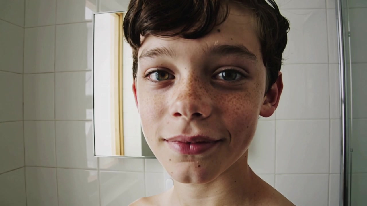 Notando la telecamera che riprende, adolescente maschio a torso nudo che si gira di profilo a viso pieno in un bagno piastrellato, specchio
