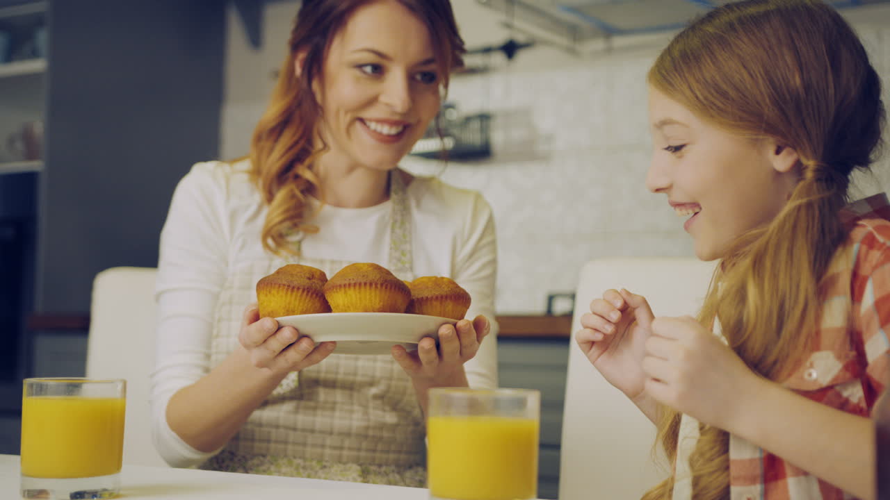 hermosa madre en el delantal trayendo y poniendo en la mesa de la cocina muffins en el plato y su adorable hija abrazándola. madre besando a una chica en la frente. en el interior