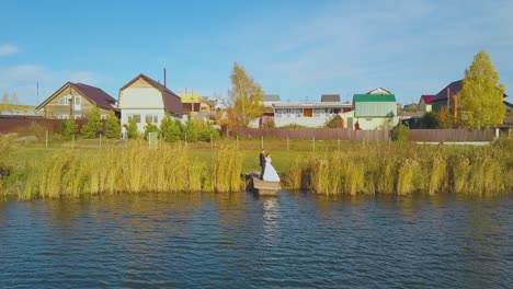 groom-and-bride-on-river-pier-at-cottage-village-aerial-view