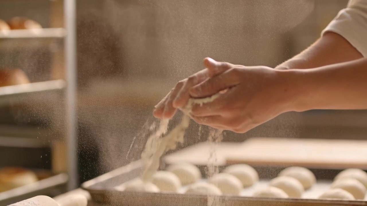 Clapping baker's hands wearing light sleeves dusting proofed rounds on bench misting flour to bake