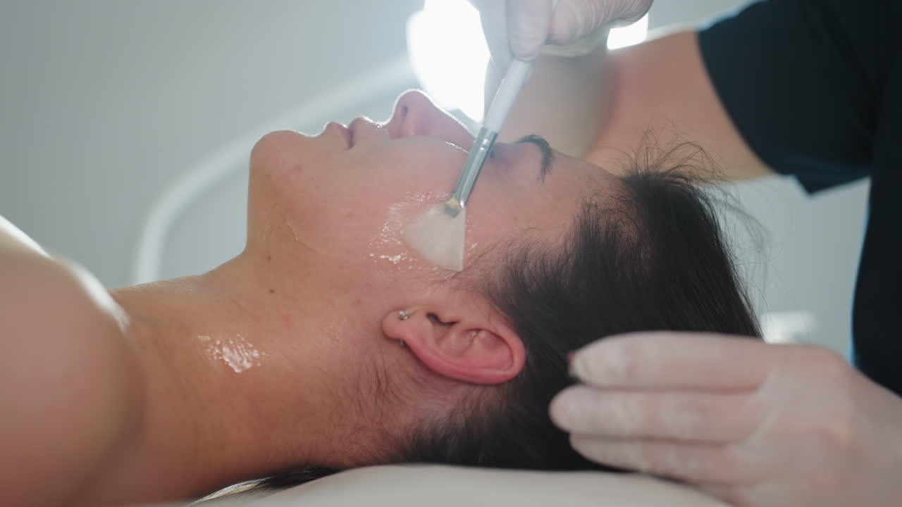 Side view of spa model lying relaxed under treatment light while beauty technician in gloves smooths translucent face gel over cheeks and forehead with brush during rejuvenating facial session