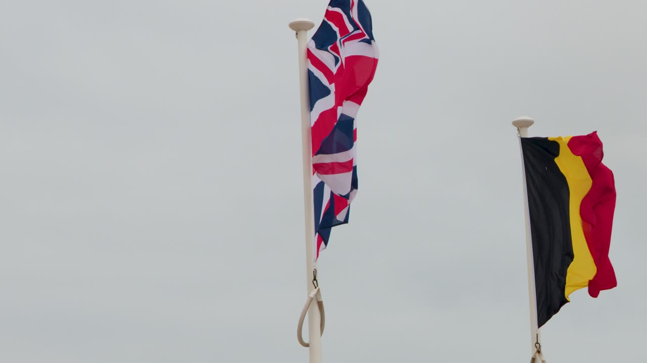 Multiple national and European flags wave vigorously on flagpoles under overcast, windy seaside conditions