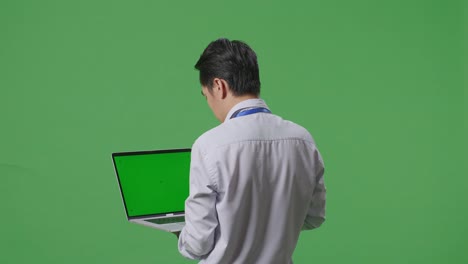 노트북 컴퓨터를 사용하는 아시아 사업가의 뒷면, 녹색 스크린을 배경으로 스튜디오에서 주변을 둘러보는, 녹색 화면 모형을 가진 노트북