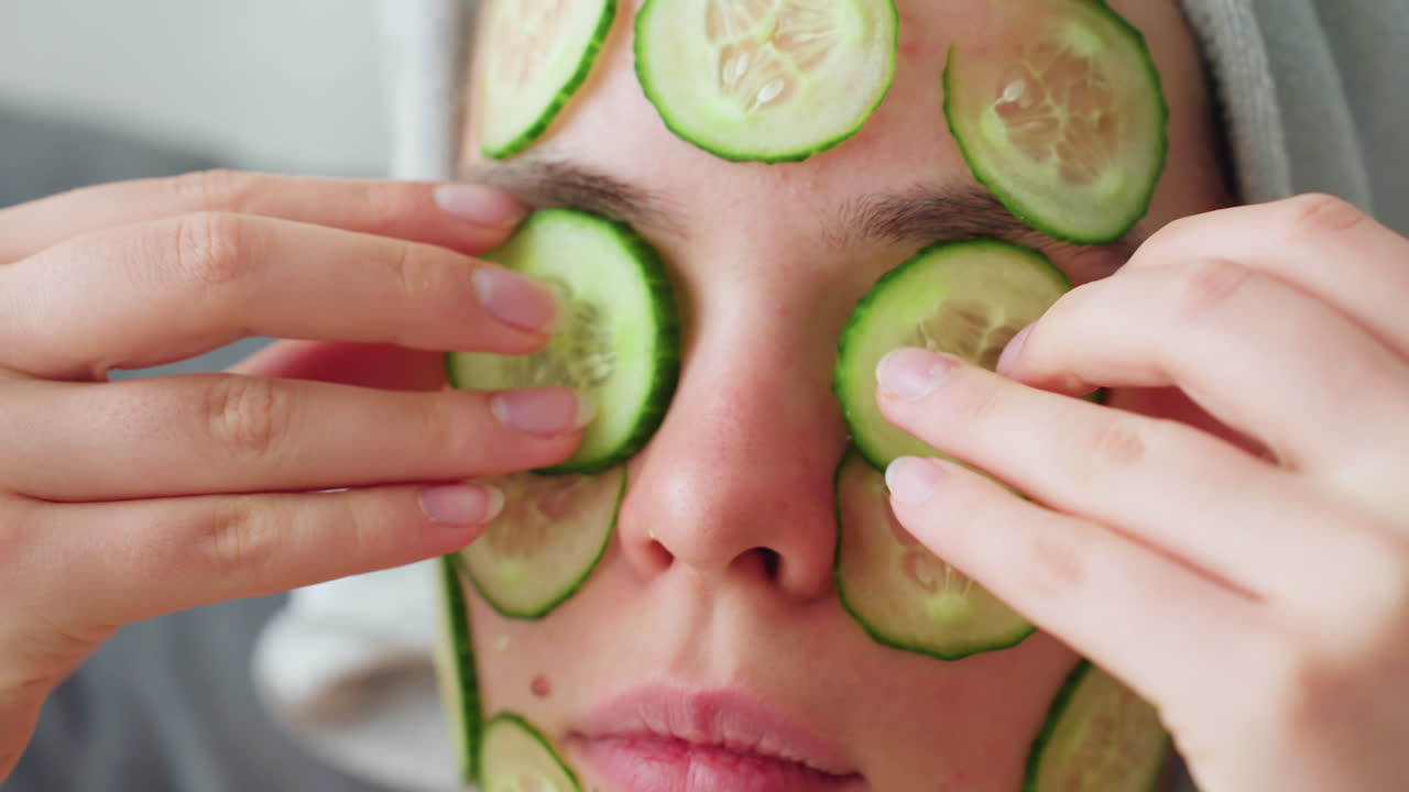 Close view of young girl placing cucumber slices on her face and eyes with both hands, she is in a relaxed state with cucumber mask for skincare. Peaceful moment of self-care